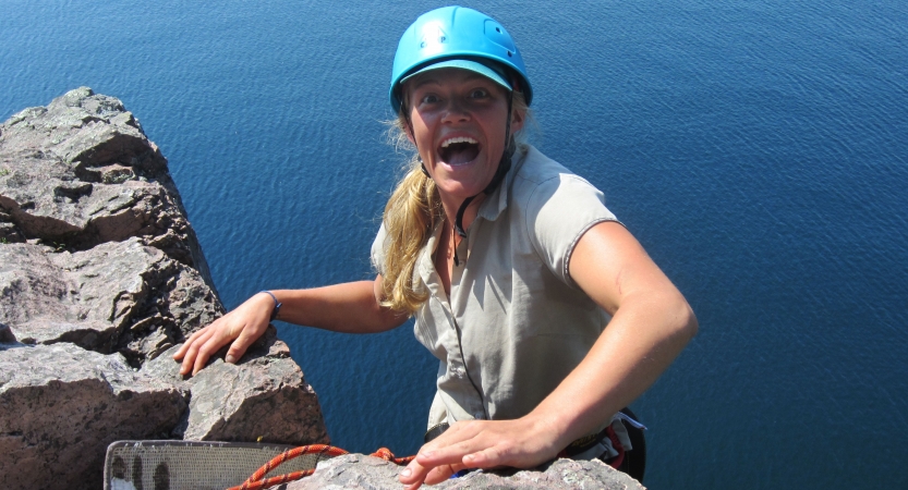 A person wearing safety gear is secured by ropes as they stand on the edge of a cliff high above a blue body of water and smile excitedly. 